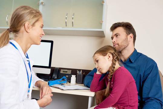 Portrait of smiling young pediatrician talking to man about health of his  little girl, all sitting at desk in office