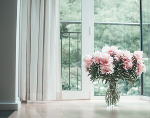 Glorious pastel pink bouquet of peonies in glass jug on  floor by window.  Flowers in interior design. Cozy home.