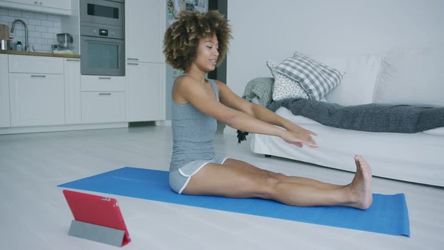 Pretty ethnic model in gray shorts sitting on mat at home watching video on tablet and practicing yoga stretching legs.
