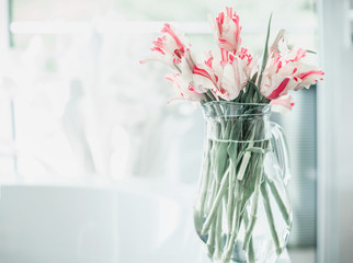 Pretty bunch of tulips in glass jug on white table at window. Flowers in interior design. Cozy home. Springtime