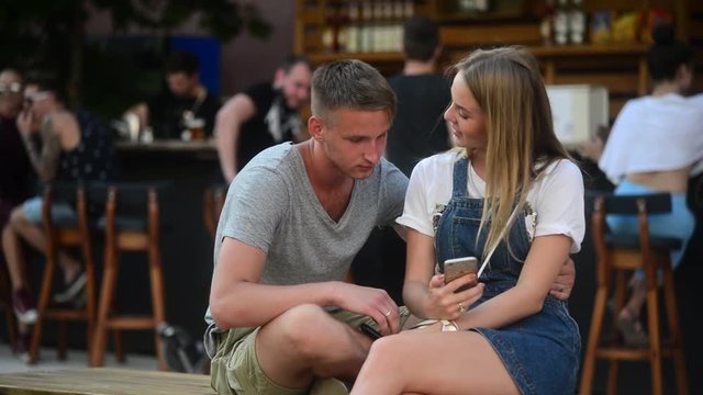 Young Guy and Girl Hug and Kiss each other sitting have fun with iphone Mobile