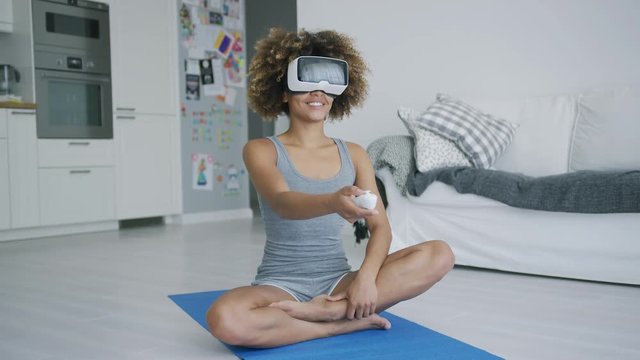 Charming ethnic woman in sportswear sitting on mat at home wearing VR goggles and enjoying another reality while doing sports.