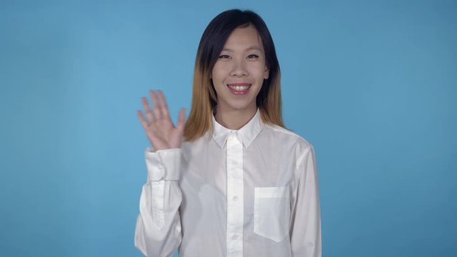 young asian woman posing showing hand gesture hello on blue background in studio. attractive millennial girl wearing white casual shirt looking at the camera smiling