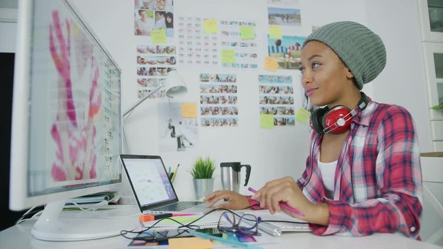 Young trendy model in hat wearing headphones and posing at working desktop with gadgets while working with computer in office.