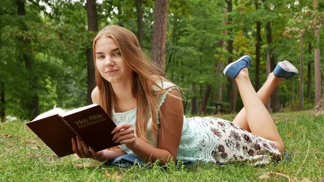 4k. Laying slender girl  read Bible in park and smile. Windy day 