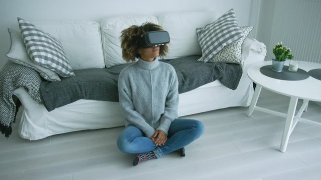 Young ethnic model in sweater sitting on floor of modern living room and wearing goggles of virtual reality experiencing another world.