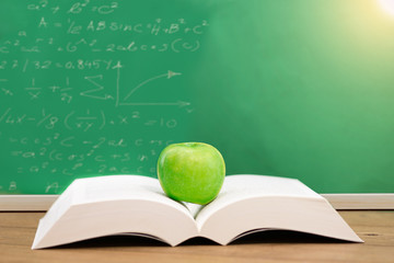 Open book with green apple on wooden table in front of blackboard