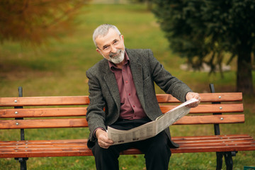 A nice grandfather with a beautiful beard in a gray jacket sits on a bench in the park and reads a newspaper