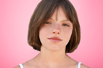 Close up portrait of freckled stylish small kid looks with narrow dark eyes directly into camera, has serious expression, isolated over pink studio background. Children, beauty, lifestyle concept