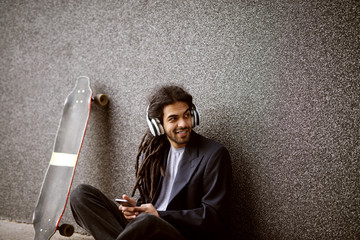 Close up of stylish playful smiling dreadlock skater man sitting leaned on the grey wall in a suit and listening music while holding a mobile and looking far away. © Dusan Petkovic