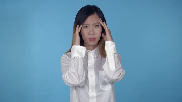 young asian happy woman posing has headache or migraine on blue background in studio. attractive millennial girl wearing white casual shirt looking at the camera