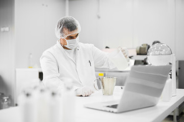 Modern professional scientist sitting at the table and pouring liquid from one flask to another at cosmetic fabric laboratory.