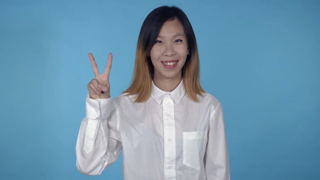 young asian woman posing showing hand gesture victory on blue background in studio. attractive millennial girl wearing white casual shirt looking at the camera smiling