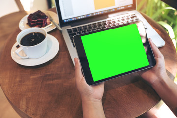 Mockup image of hands holding black tablet pc with blank green screen , laptop , coffee cup and cake on wooden table in cafe