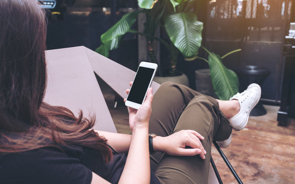 Mockup image of woman's hands holding white mobile phone with blank black screen on thigh while sitting and chilling in modern cafe