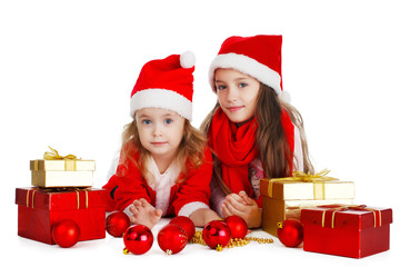 Two little girls in hats of Santa Claus lying down with gifts. White background