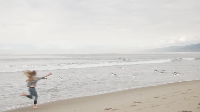 teen girl run on Santa Monica beach and seagulls flying from ground in cloudy november day shot with gimbal