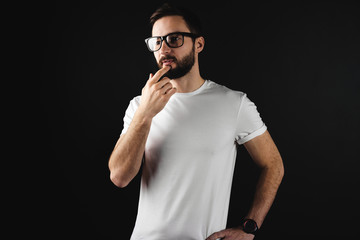 Pensive bearded guy in glasses wearing in white blank t-shirt on black background