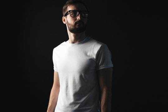 Serious Bearded Man In Glasses Wearning In White T-shirt. Hipster In Shadows In A Blank Tshirt On Dark Background