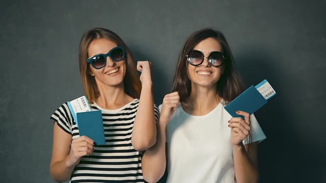 Thrilled girls with tickets and passports smiling and showing tumbs up in the grey background
