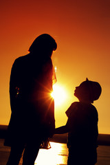 Silhouettes of mom and son holding hands and admiring the beautiful view of the sea on sunny evening at sunset time