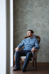 Portrait of bearded male, dressed in a plaid shirt sits on a chair over dark grey background.