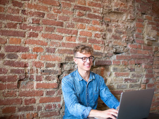 Young man freelancer working on his web site on portable laptop computer, sitting in modern interior against brick wall. Stylish hipster guy working in internet via notebook. Student using net-book