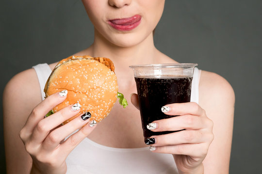 Young woman eating hamburger and cola on gray background. Junk food and fast food concept