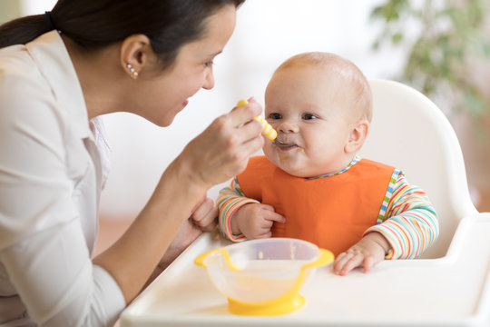Food, child and parenthood concept - mom with puree and spoon feeding baby at home