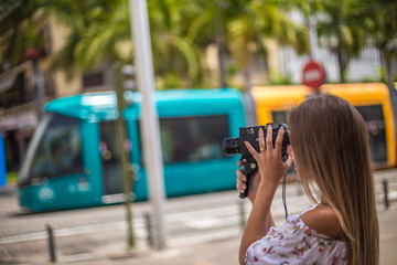 Tenerife. Spain. Cute blonde girl is observing and  filming streets in Santa Cruz de Tenerife with colourful tram on the background. Girl travels concept. Travel to Canary Islands.