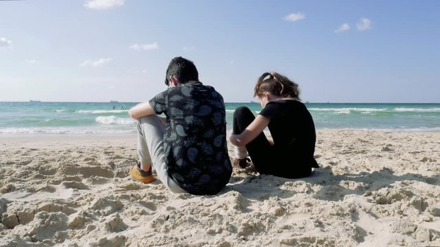 Teenagers, girl and guy are sitting on the beach and talking.