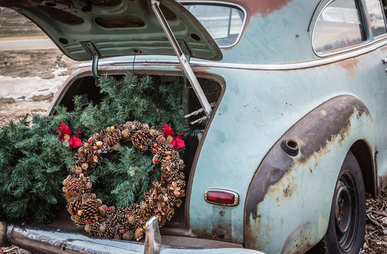 horizontal close up rustic image of the back end of an old green vintage car with the trunk open and a brown acorn Christmas wreath and Christmas tree in the back of trunk.