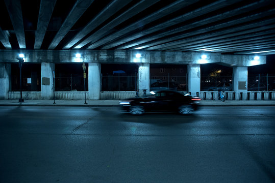 Gritty Dark Chicago Highway Bridge And City Street With A Passing Black Car And Bicycle Stand At Night.