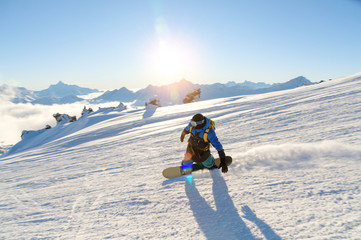 A snowboarder in a ski mask and a backpack is riding on a snow-covered slope leaving behind a snow...