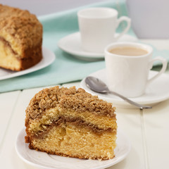 Close-up piece of homemade Cinnamon crumble coffee cake and cup of milk tea. White wooden background. Square photo.