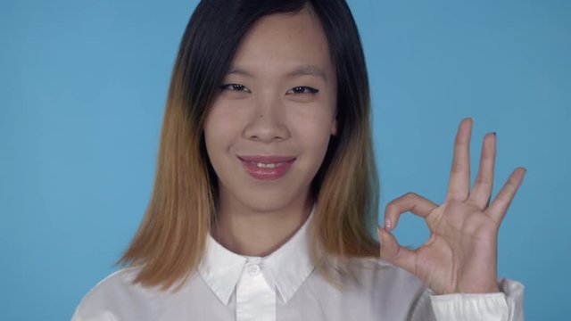 close up portrait young asian woman laughing shows sign symbol ok on blue background in studio. attractive millennial girl looking at the camera smiling