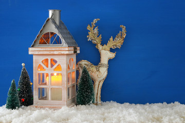Wooden old house with candle, white deer next to christmas trees over the snow and blue nackground.