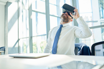 Portrait of mature bearded businessman using VR glasses sitting at table in office taking break from work, copy space