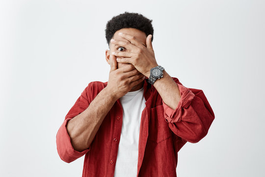 Close Up Of Young Dark-skinned Man With Afro Hairstyle In White E-shirt Under Red Shirt Clothing Face With Hands, Looking Through Fingers With Scared Face Expressions.