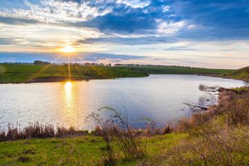 Colorful landscape on the river bank in spring, orange sun hiding behind blue clouds in a beautiful sky. Sunset on the lake in Russia.