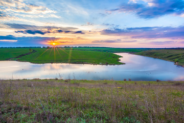 Obraz premium Colorful landscape on the river bank in spring, orange sun hiding behind blue clouds in a beautiful sky. Sunset on the lake in Russia.