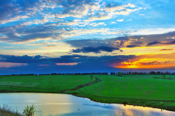 Colorful landscape on the river bank in spring, orange sun hiding behind blue clouds in a beautiful sky. Sunset on the lake in Russia.
