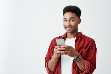 Positive emotions. Close up of young good-looking dark-skinned male with afro hairstyle in white t-shirt and red shirt smiling with teeth, chatting with friend on smartphone