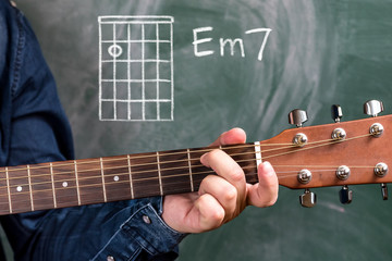 Man in a blue denim shirt playing guitar chords displayed on a blackboard, Chord Em7