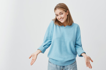 Indoor shot of beautiful Caucasian young female with straight fair hair shrugging her shoulders and smiling broadly feeling confused while refusing to go out on date with guy she doesn't like