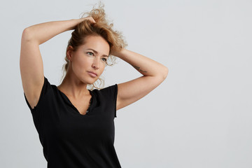 Emotional beautiful young adult woman portrait. Studio shoot. Isolated on White Background. A woman in a black T-shirt with a deep neckline lifted her hair with her hands and looks to the right.