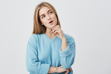 Dark-eyed young woman with fair hair wearing blue sweater having pensive expression thinking over her plans, looking aside, standing against gary background with copy space for advertisment or text