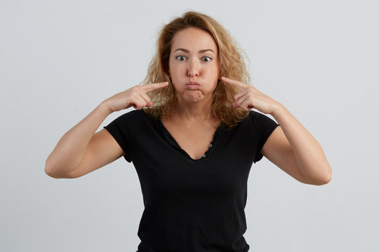 Emotional beautiful young adult woman portrait. Studio shoot. Isolated on White Background. A woman in a black T-shirt inflates her cheeks with wide open eyes. She wants to poke her fingers on her