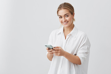 Adorable hipster Caucasian female with blonde hair checking her news feed or messaging via social networks, using free wi-fi on mobile phone, smiling, posing against gray studio wall