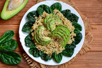 Spiced quinoa porridge. Vegan quinoa with spinach and avocado salad on a white plate. Brown wooden table. Rustic style. Top view